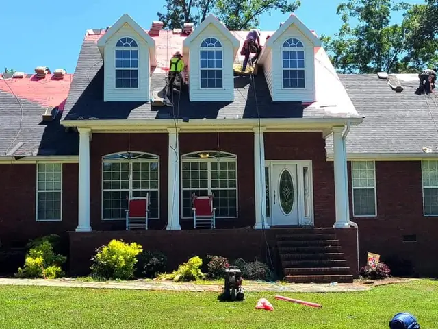 Roofers working on a brick home with three dormers, red underlayment on part of the roof and dark shingles on the rest