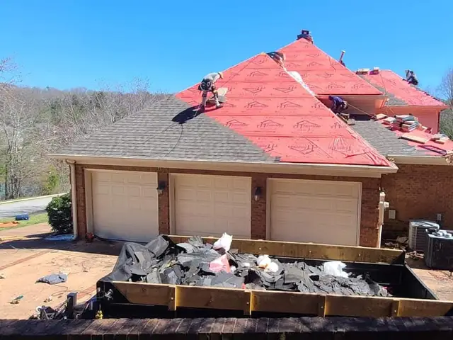 Roofers on a hip roof covered in red underlayment above a three-car garage, with a dumpster of old shingles below