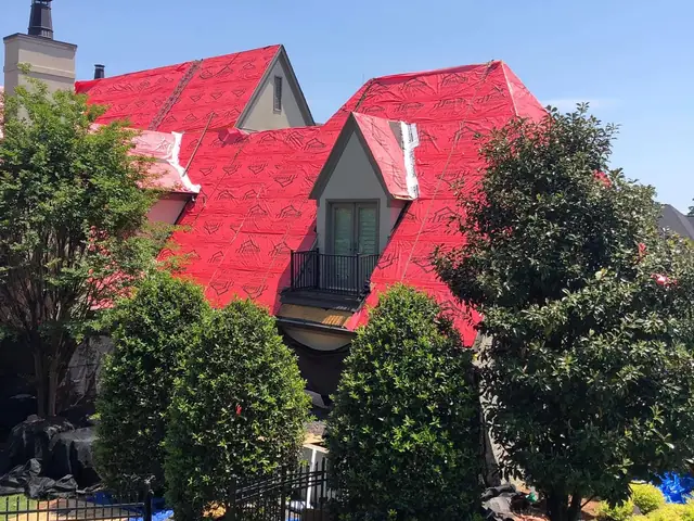 A large home with red underlayment laid across its roof, seen through trees