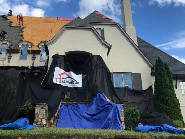 Roofers on a large stucco home mid-replacement, with a company banner hung on the safety netting